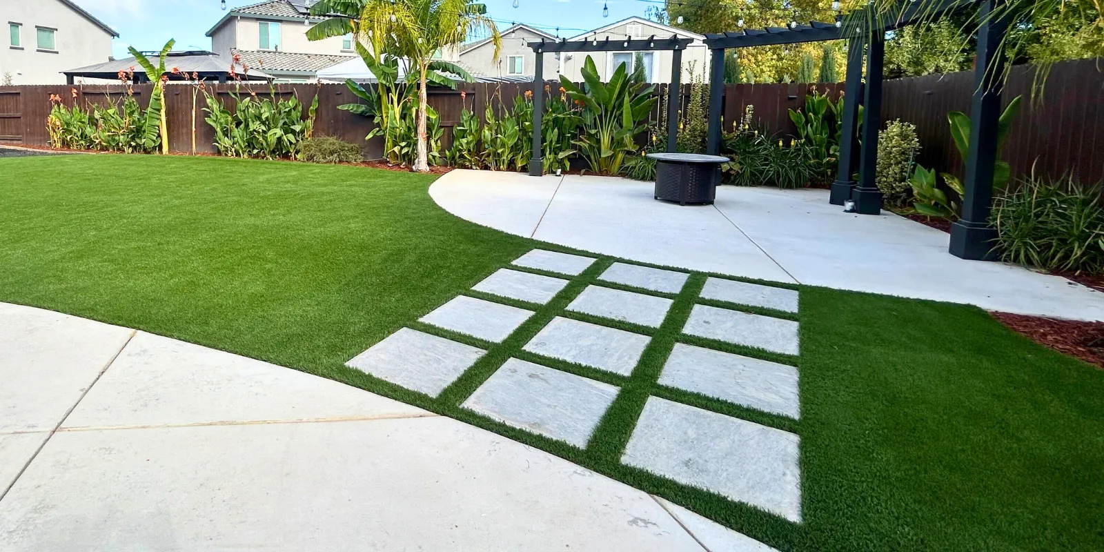 Modern backyard with artificial grass lawn, concrete patio, stone paver walkway, and pergola surrounded by tropical plants and palm trees. Lush artificial turf lawn with decorative stone pavers