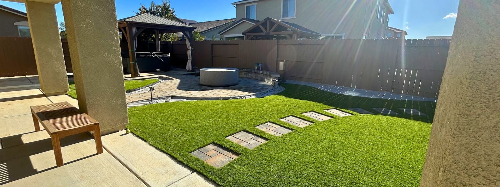 Modern backyard with artificial grass, stone paver walkway, fire pit area, and wooden gazebo creating a cozy outdoor living space. Backyard featuring lush artificial turf, stone pathway, and gazebo