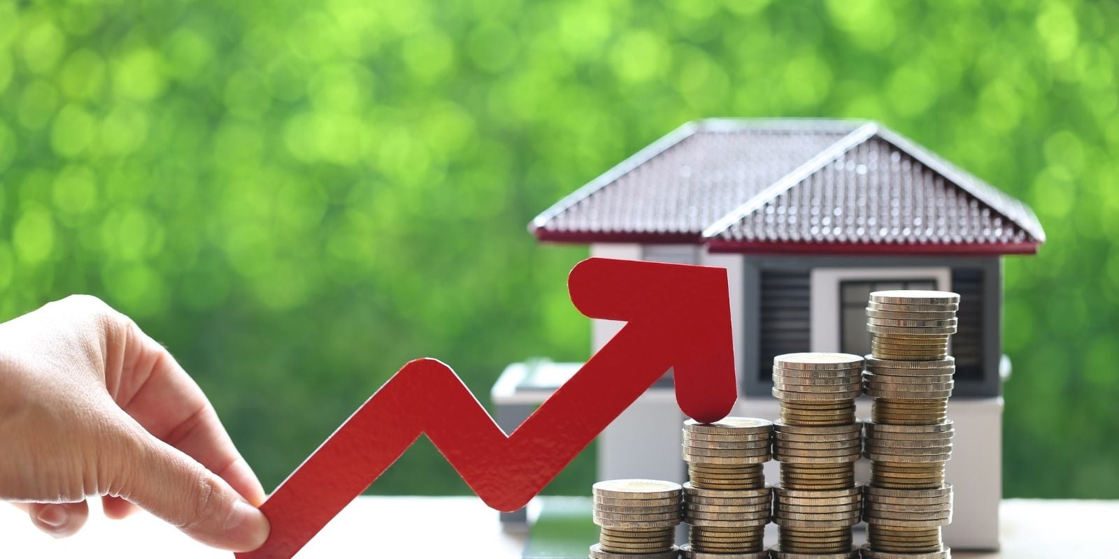 Hand holding a red upward arrow next to stacks of coins and a small house model, symbolizing rising home values, real estate investment, and property market growth. stacks of coins