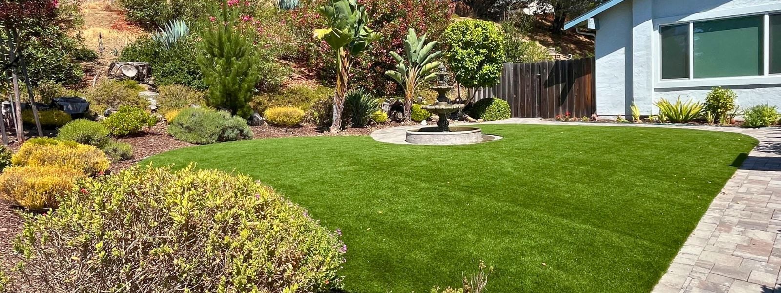 Beautiful residential front yard with lush artificial grass lawn, landscaped shrubs, and a decorative fountain near a modern home. Lush green artificial turf lawn surrounded by colorful shrubs