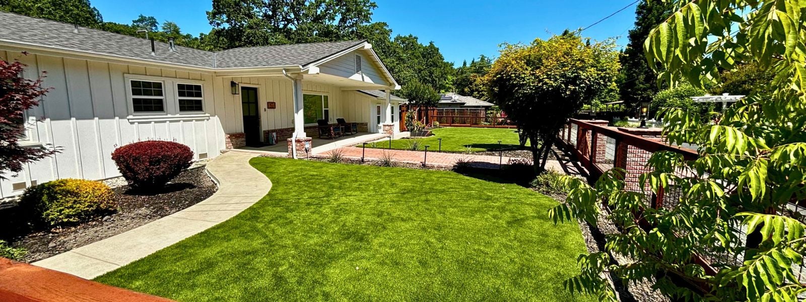 Beautiful front yard of a white ranch-style home featuring lush artificial grass, landscaped garden beds, and a concrete pathway leading to the entrance. Lush artificial turf lawn enhancing curb appeal of a San Jose home